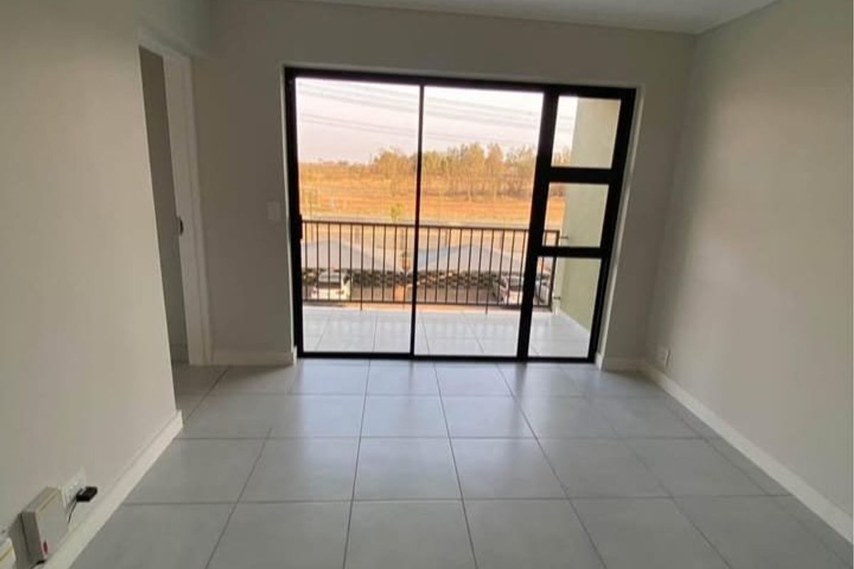 A bright, empty living area featuring neutral tiled flooring and a large sliding glass door providing access to a private balcony.