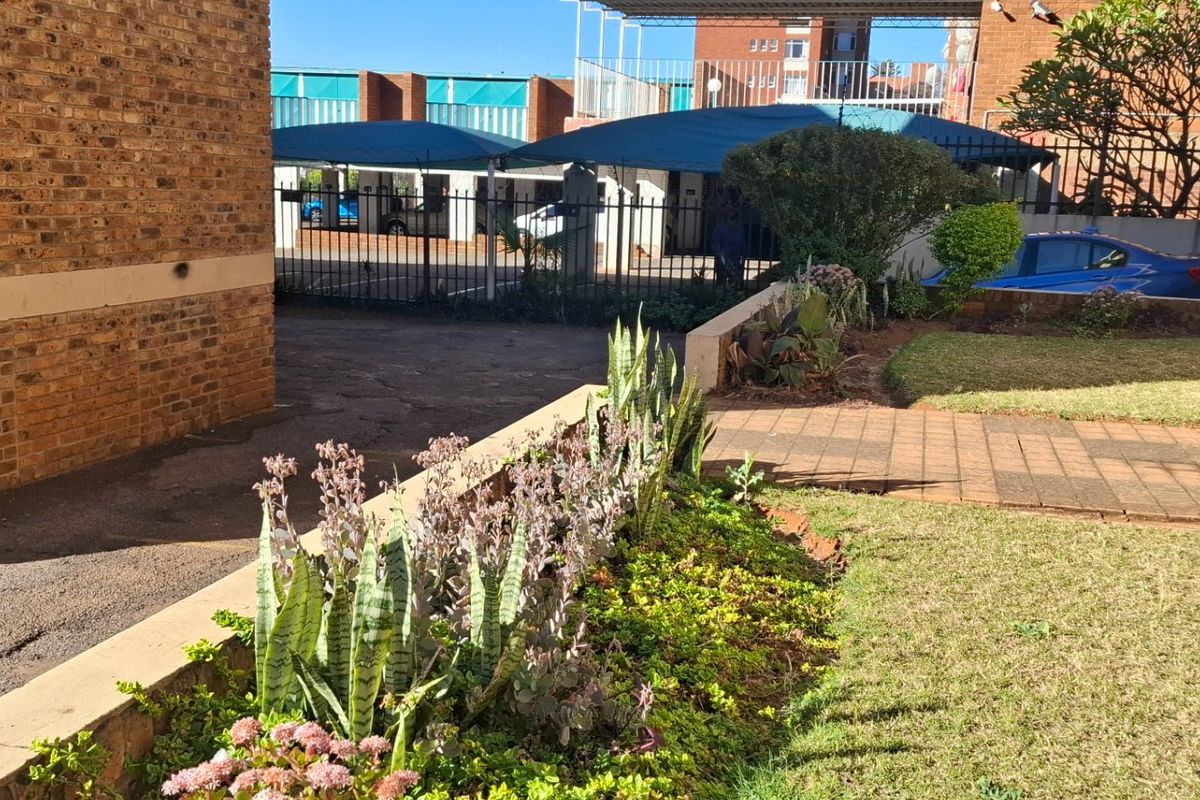 A view of the apartment complex exterior featuring a landscaped garden, paved pathways, and a brick facade with secure perimeter fencing and carport parking.