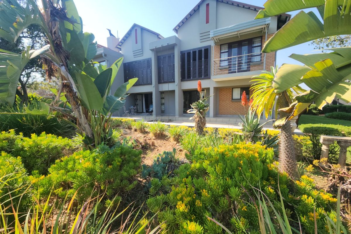 Rear view of a multi-story residential property featuring a paved patio, upper-level balcony, and dark window shutters, set against a landscaped garden.