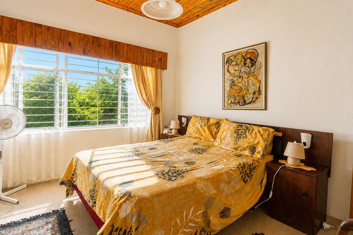 A bright bedroom featuring a timber ceiling, built-in headboard, and large window with security bars, offering a comfortable and functional living space.