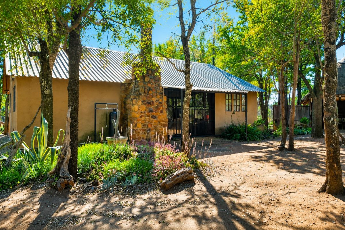 A charming rustic cottage featuring a stone chimney, corrugated metal roof, and secure entrance, set within a tranquil, tree-lined garden environment.