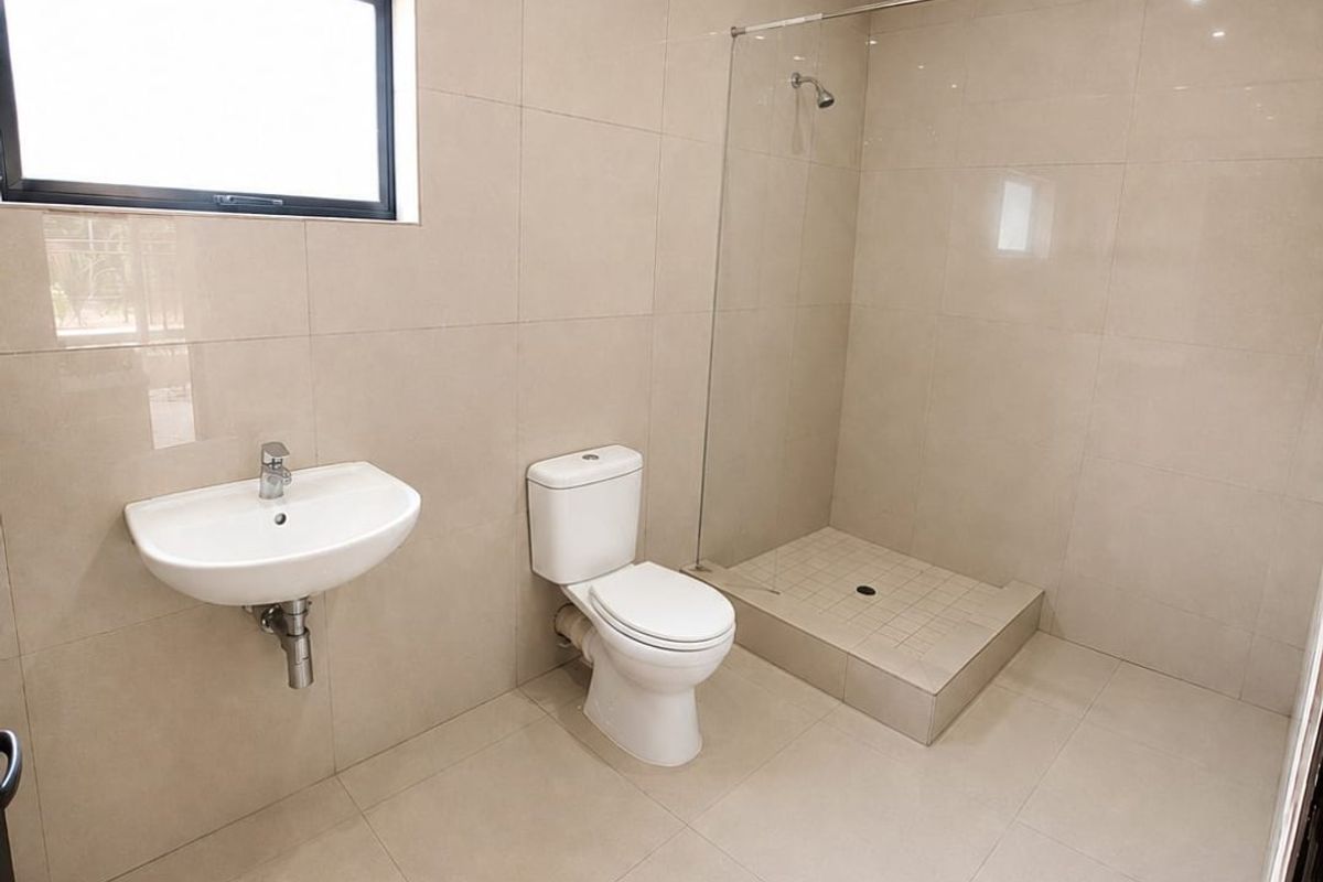 A modern, clean bathroom featuring floor-to-ceiling beige tiling, a wall-mounted basin, a white toilet, and a walk-in shower with a glass screen.