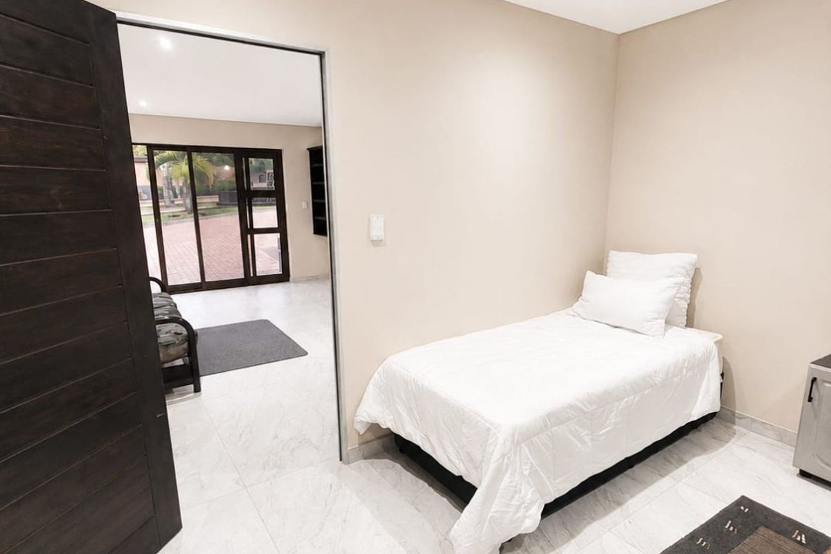 A clean, neutral-toned bedroom featuring modern tiled flooring, recessed downlighting, and a solid wood door, with direct access to an adjacent living area.