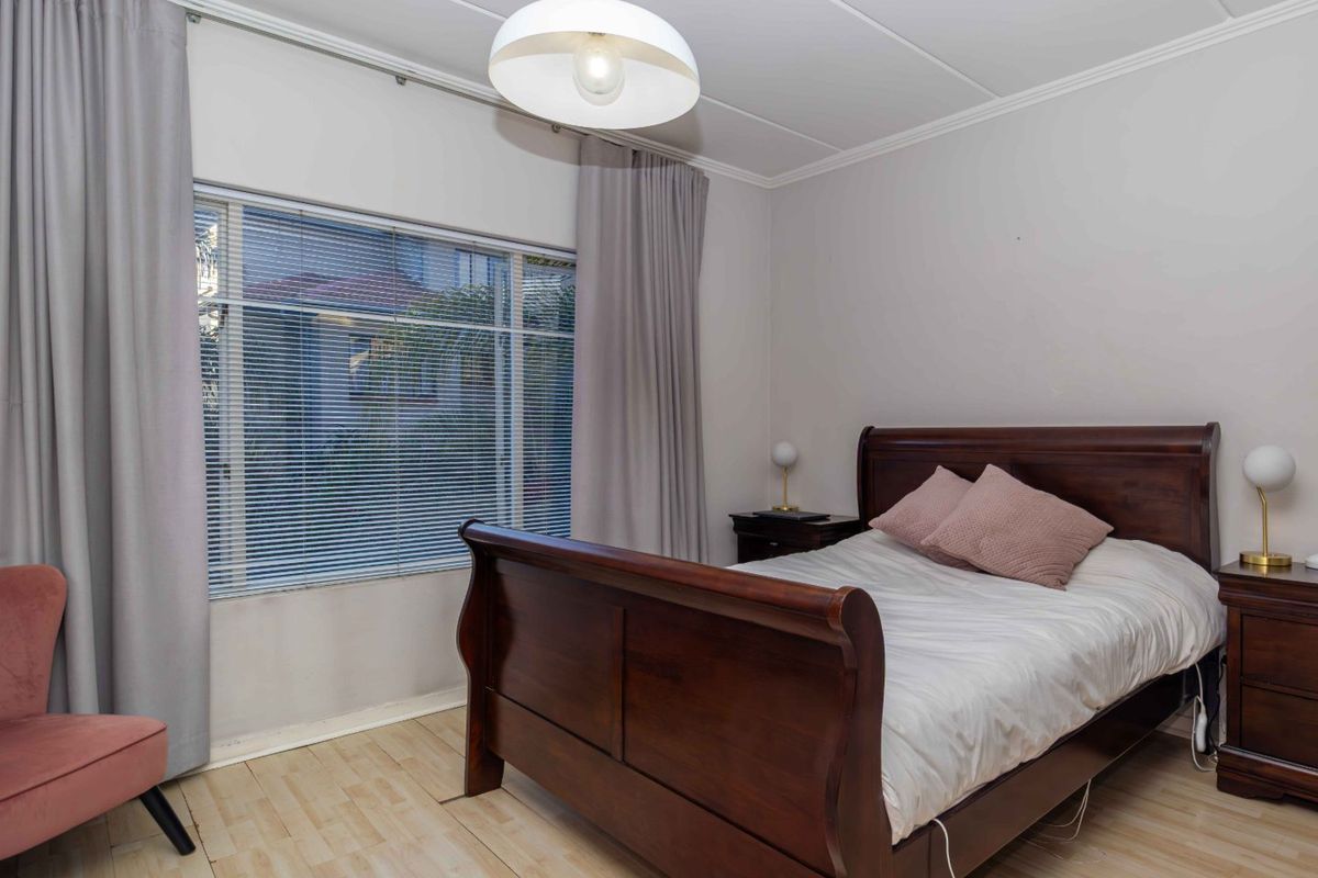 A well-lit bedroom featuring laminate flooring, neutral walls, and large windows with venetian blinds, offering a comfortable and functional living space.