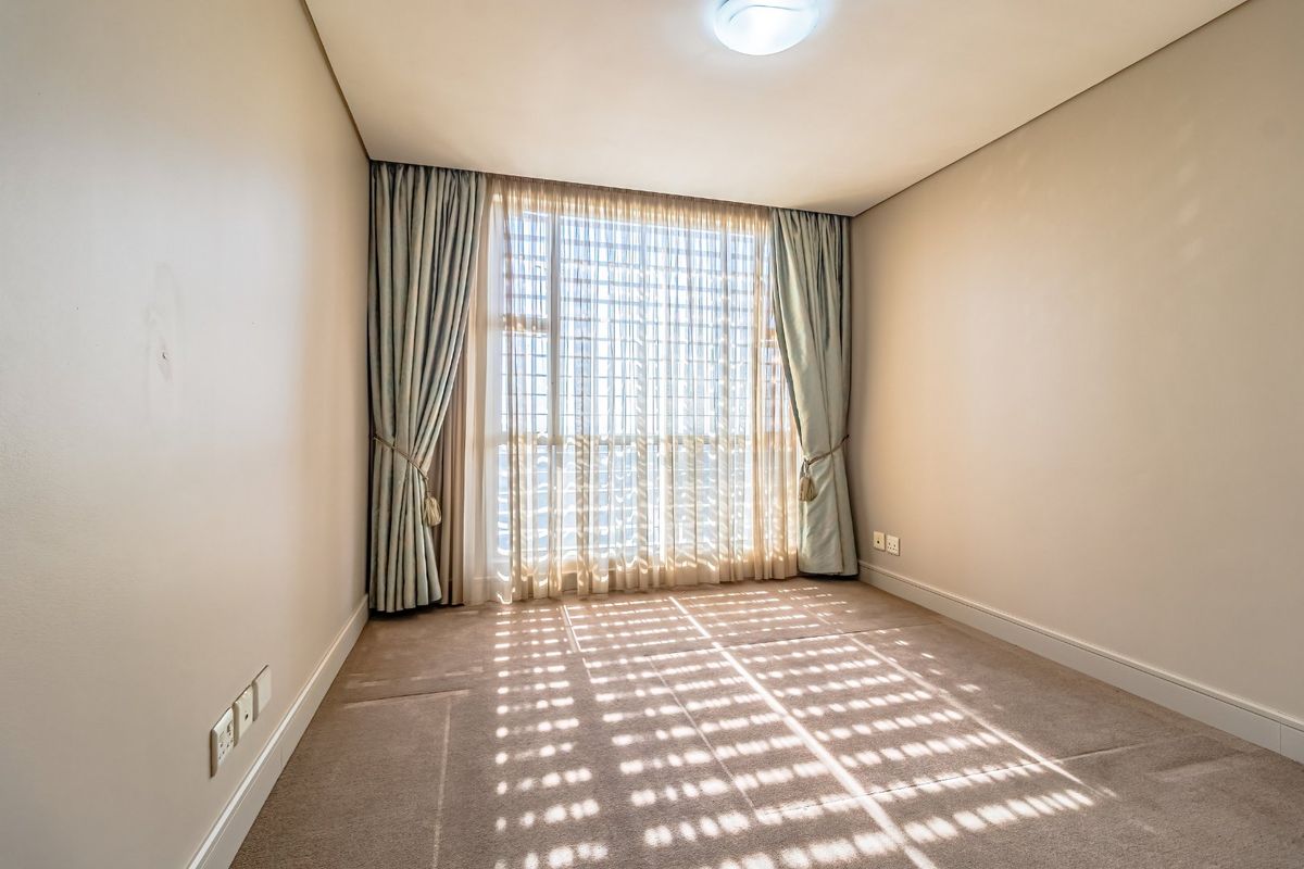 A bright, empty bedroom featuring neutral carpet flooring, wall-mounted electrical outlets, and floor-to-ceiling window treatments.