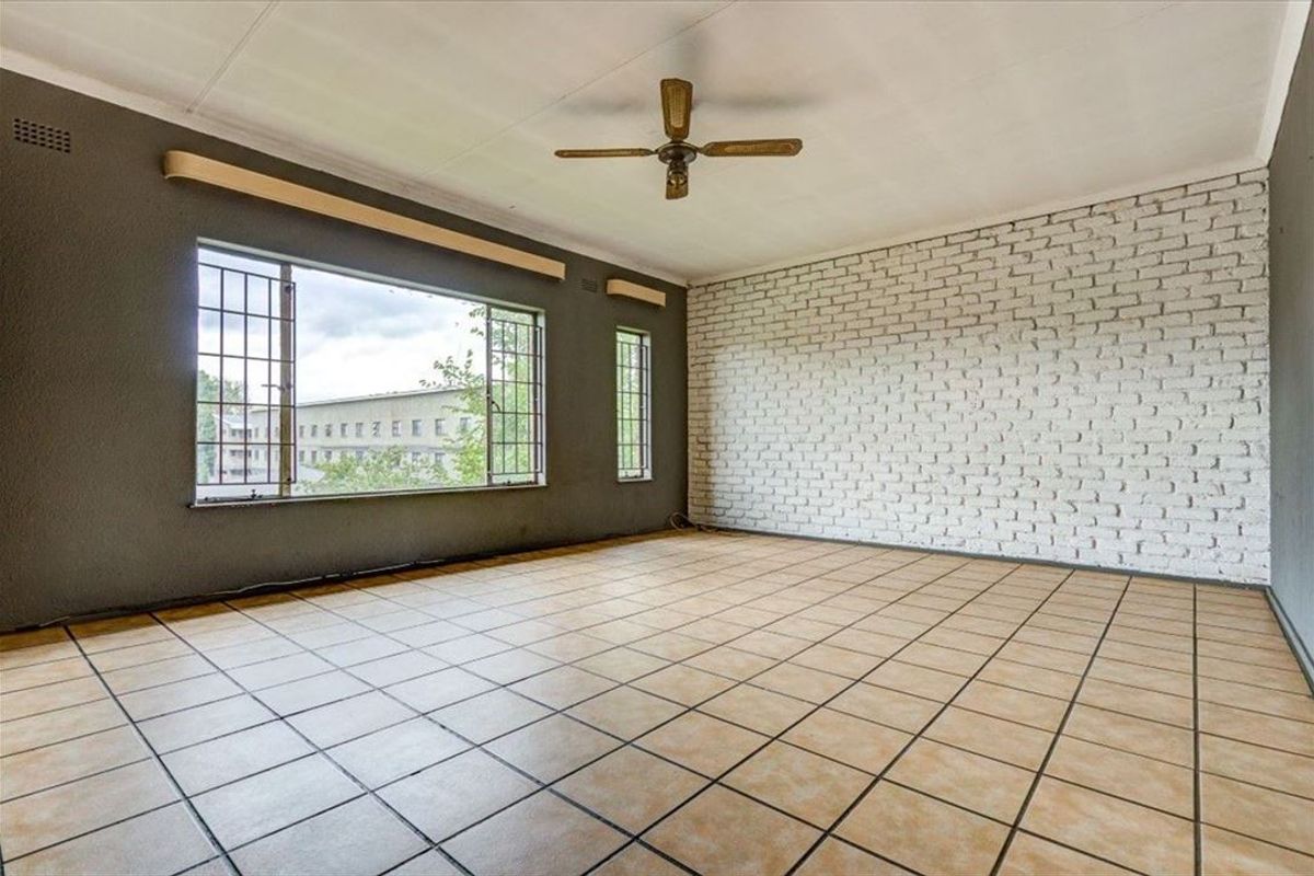 Spacious living area featuring tiled flooring, a ceiling fan, and a distinctive white-painted exposed brick feature wall with large windows.
