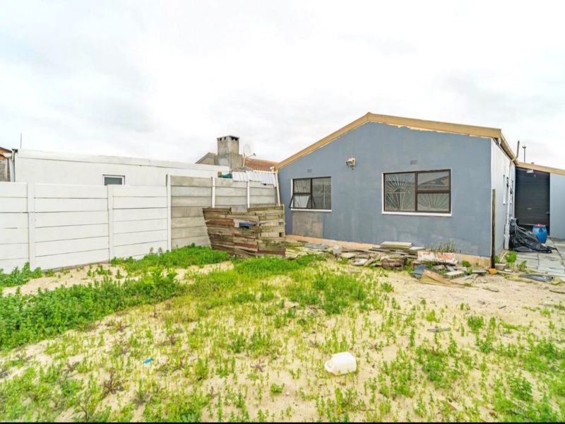 Rear view of the property featuring a large sandy yard, precast concrete boundary walls, and a single-story structure with aluminium window frames.