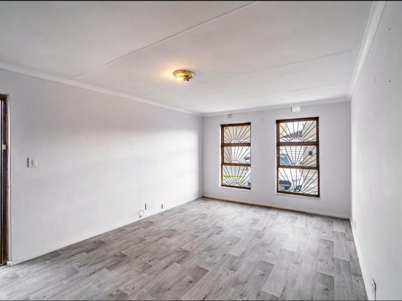 An empty living room featuring modern grey laminate flooring, white walls, and windows fitted with decorative security bars.