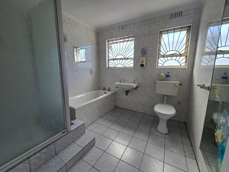 A functional, tiled bathroom featuring a bathtub, separate shower enclosure, wall-mounted basin, and toilet, with security bars installed on the windows.