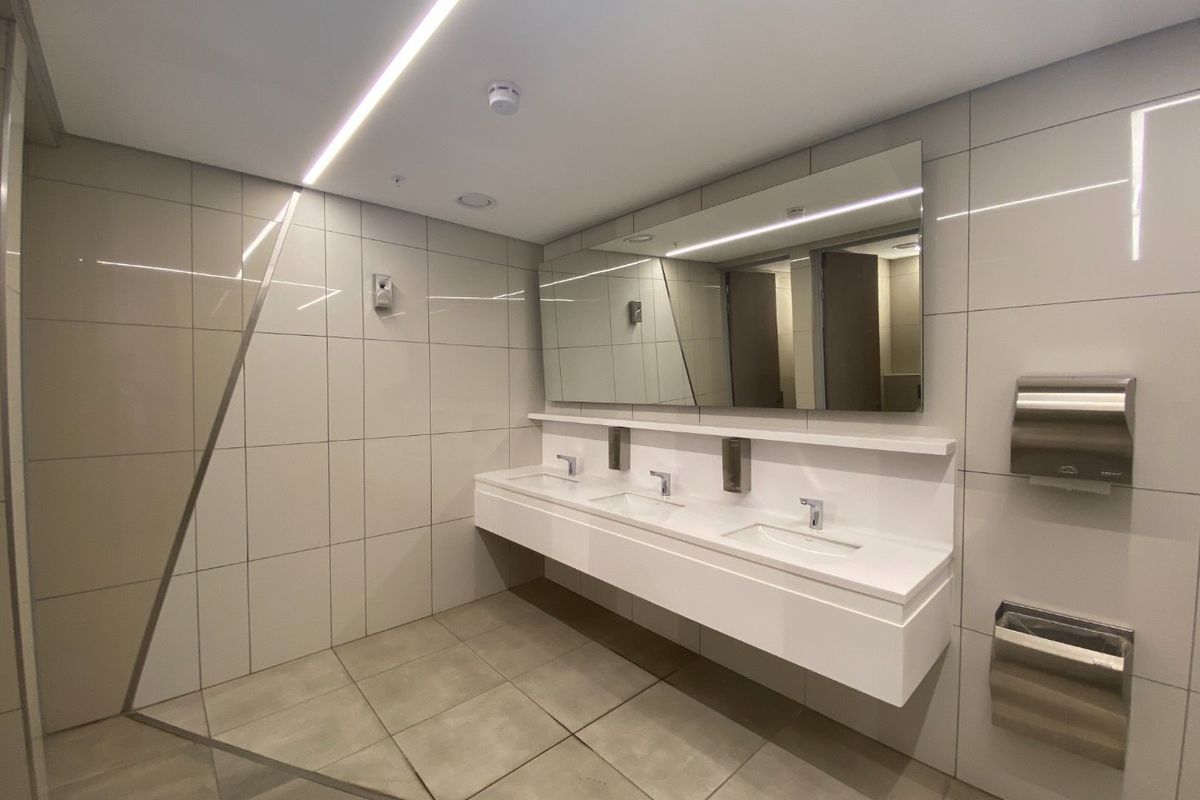Modern commercial bathroom featuring a sleek triple vanity with integrated sinks, large mirror, recessed lighting, and high-quality tiled finishes.
