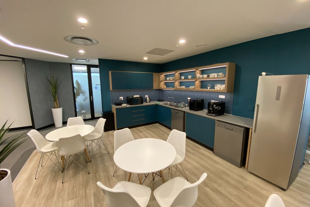 Modern commercial kitchenette featuring teal cabinetry, subway tile backsplash, integrated shelving, and recessed lighting, providing a functional break area for office staff.