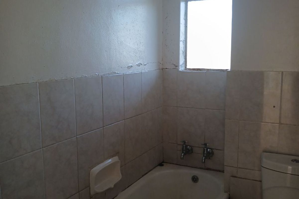 A dated bathroom featuring a standard bathtub, toilet, and tiled finishes, requiring cosmetic updates and maintenance to the wall surfaces and window frame.