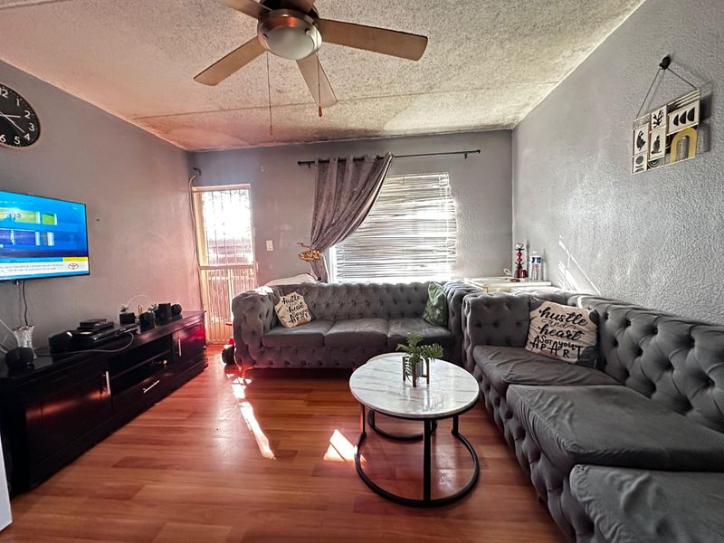 Living room featuring laminate flooring, a ceiling fan, and security gate access. The ceiling shows visible signs of water damage and peeling paint.