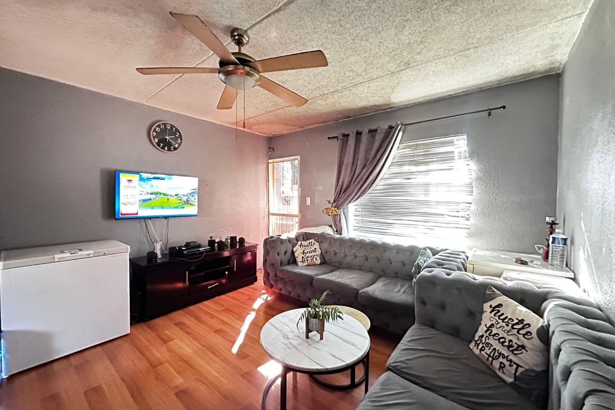A living room featuring laminate flooring, a ceiling fan, and natural light through window blinds, with a security gate at the entrance.