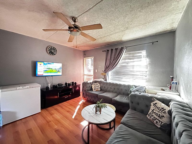 A living room featuring laminate flooring, a ceiling fan, and natural light through window blinds, with a security gate at the entrance.