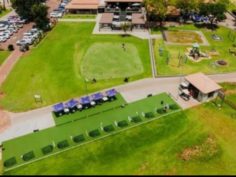 Aerial view of a well-maintained golf driving range facility featuring manicured greens, paved pathways, and associated clubhouse infrastructure within a residential estate.