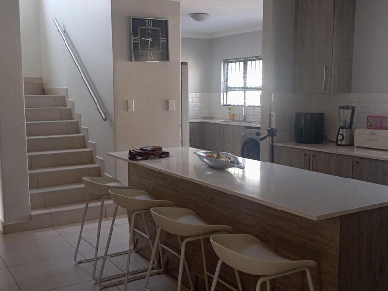Open plan kitchen featuring a central island with stone countertop, light wood cabinetry, and tiled flooring, adjacent to a staircase.