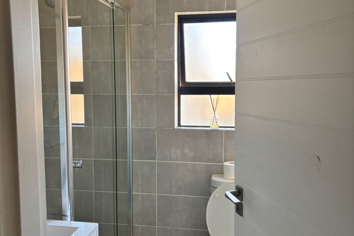 A compact bathroom featuring a glass-enclosed shower, a modern vanity, and a toilet, finished with neutral wall and floor tiling.