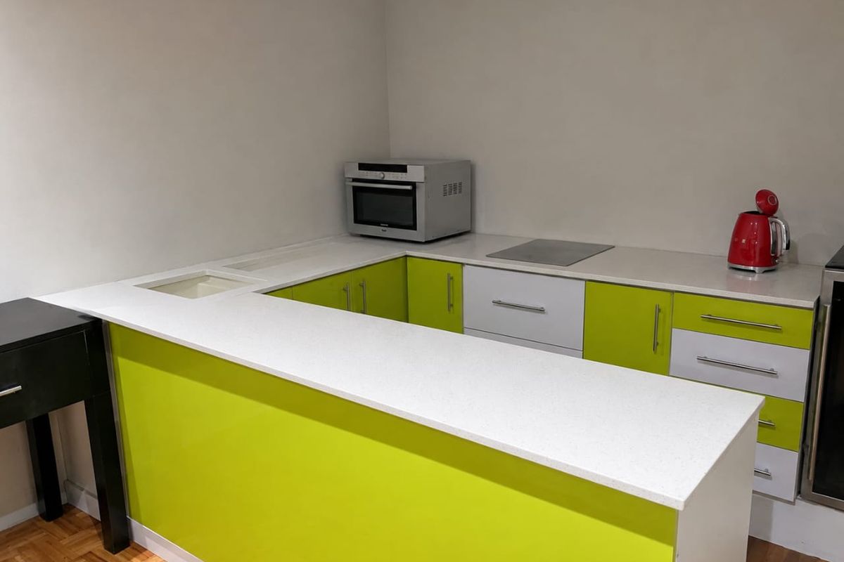 A modern kitchen featuring vibrant lime green cabinetry, white stone countertops, integrated cooktop, and built-in microwave, set against a neutral wall with parquet flooring.
