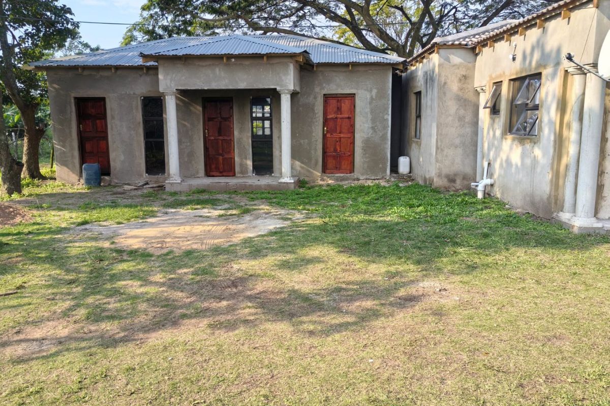 Exterior view of a commercial building featuring multiple entrance doors, concrete columns, and an unfinished plaster facade set on a large grassed plot.