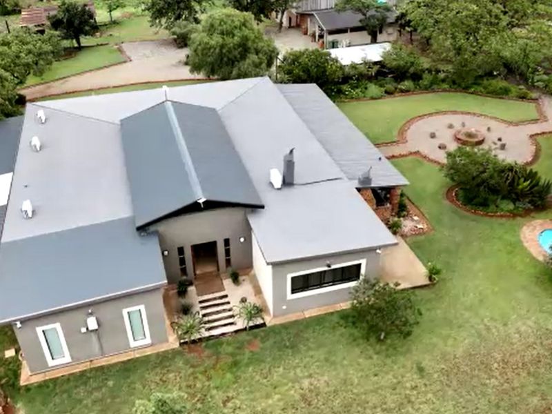 Aerial view of a modern farmhouse featuring a grey metal roof, spacious garden, swimming pool, and paved outdoor entertainment area.