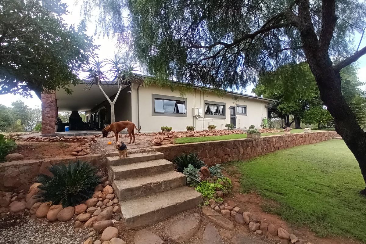 A rear view of the farmhouse featuring a covered patio, stone retaining wall, landscaped garden, and mature trees.