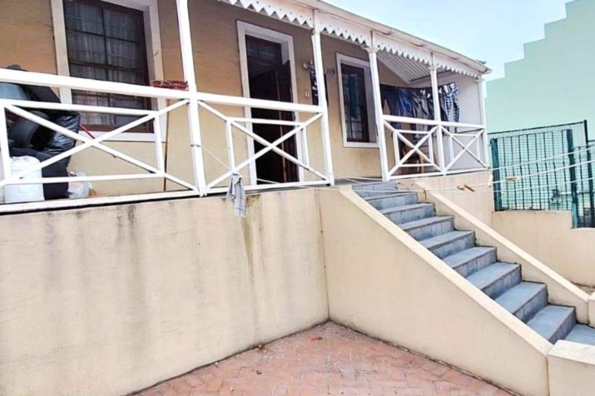 Exterior view of a raised patio area with white railings, concrete stairs, and paved ground level, showing signs of weathering and maintenance needs.