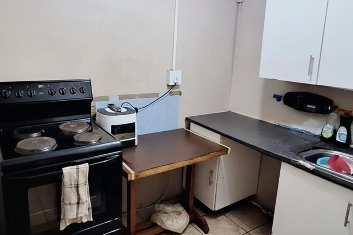 A compact kitchen area featuring a freestanding stove, dark laminate countertops, a stainless steel sink, and white cabinetry with visible wear.