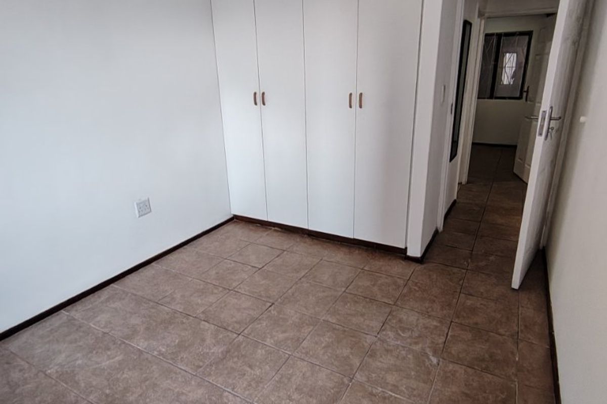 A clean, empty bedroom featuring tiled flooring, built-in white cabinetry, and a ceiling light fixture, with access to an adjacent hallway.