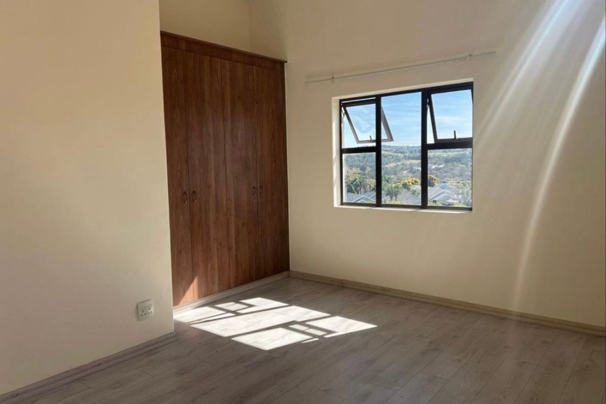 A bright, empty bedroom featuring wood-look laminate flooring, built-in wooden cupboards, and a window offering scenic elevated views.