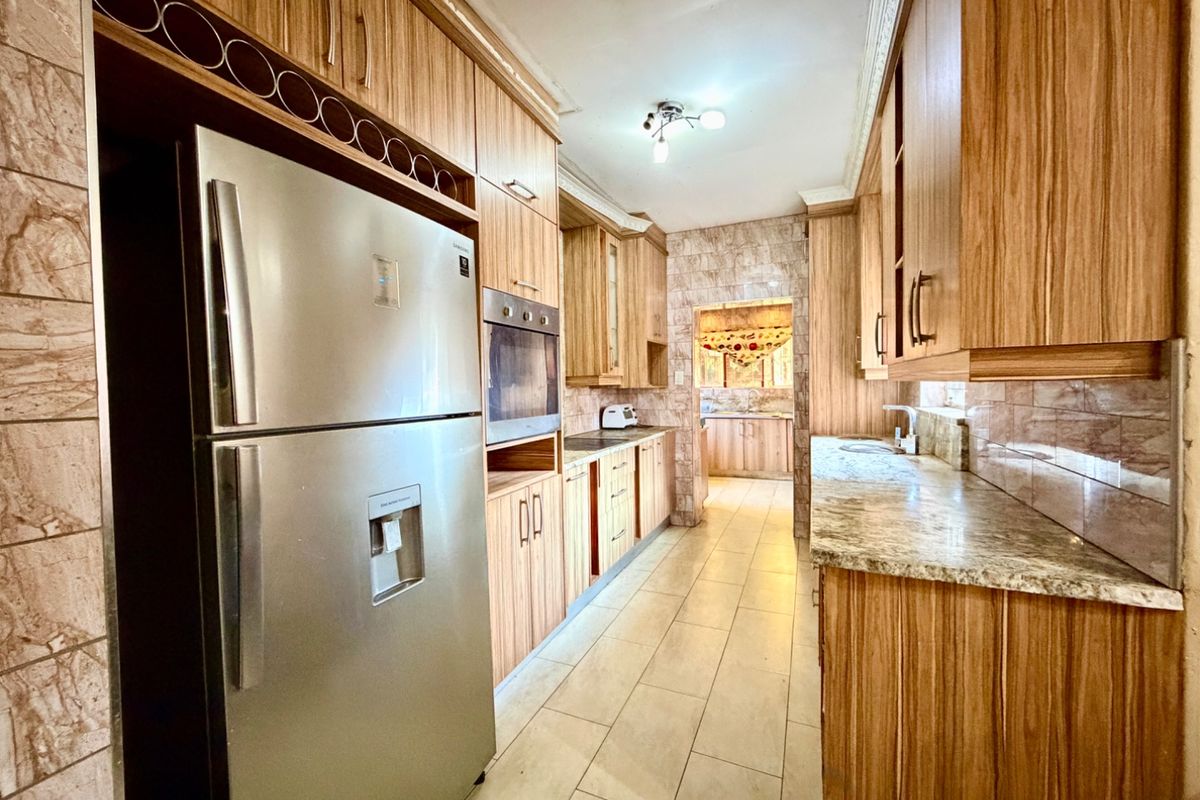 A functional kitchen featuring wood-grain cabinetry, granite countertops, tiled flooring, and a built-in oven, with an adjacent scullery area visible in the background.