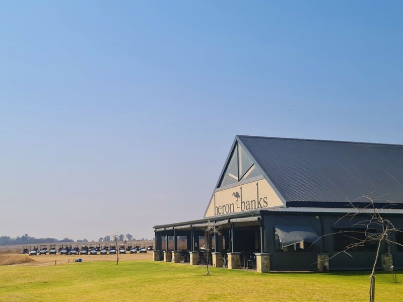 Exterior view of the Heron Banks clubhouse facility, featuring a modern pitched roof design, expansive lawn, and adjacent golf cart parking area.