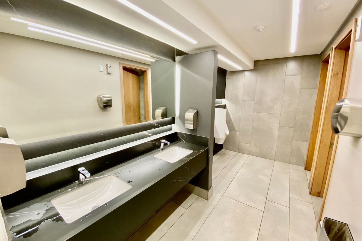 Modern commercial restroom featuring a dual-basin vanity with stone countertop, large integrated mirror, recessed LED lighting, and neutral large-format floor and wall tiling.