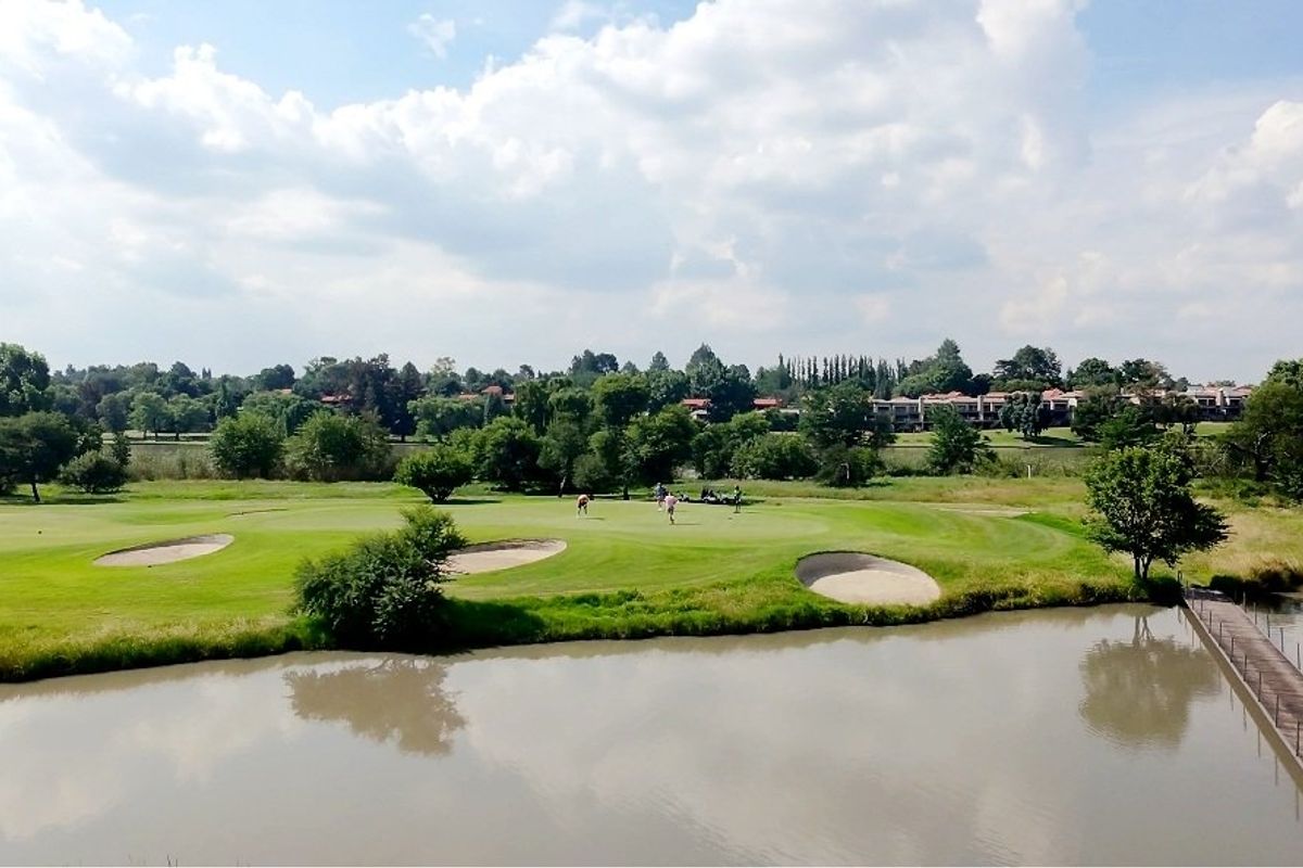 A scenic aerial view of a golf course featuring a water hazard, sand bunkers, and a wooden footbridge, set against a lush landscape.