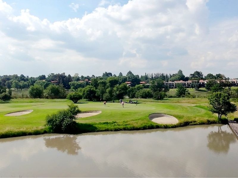 A scenic aerial view of a golf course featuring a water hazard, sand bunkers, and a wooden footbridge, set against a lush landscape.