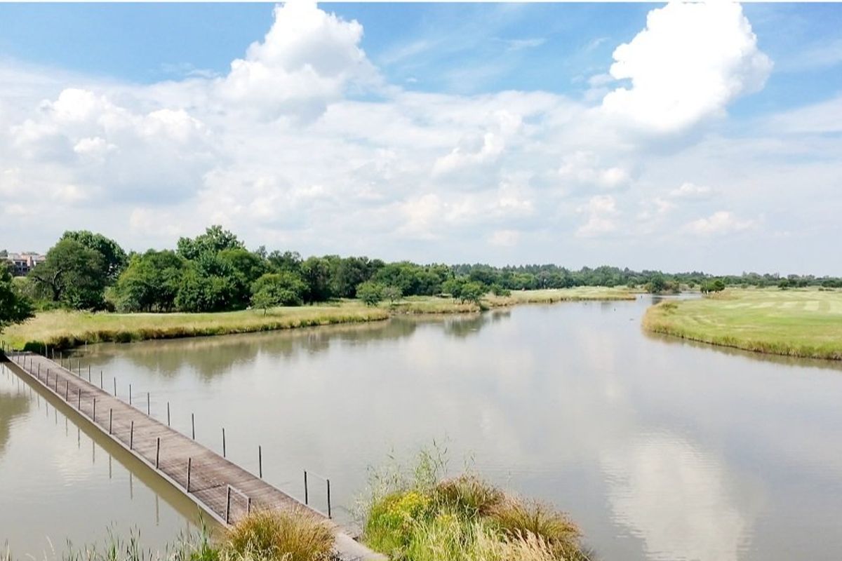A scenic view of a tranquil lake with a pedestrian footbridge, surrounded by lush green landscape and mature trees.