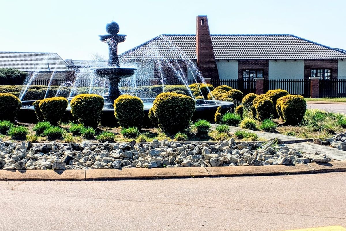 A landscaped garden area featuring a central tiered water fountain, manicured shrubs, and decorative rock borders in a residential estate setting.