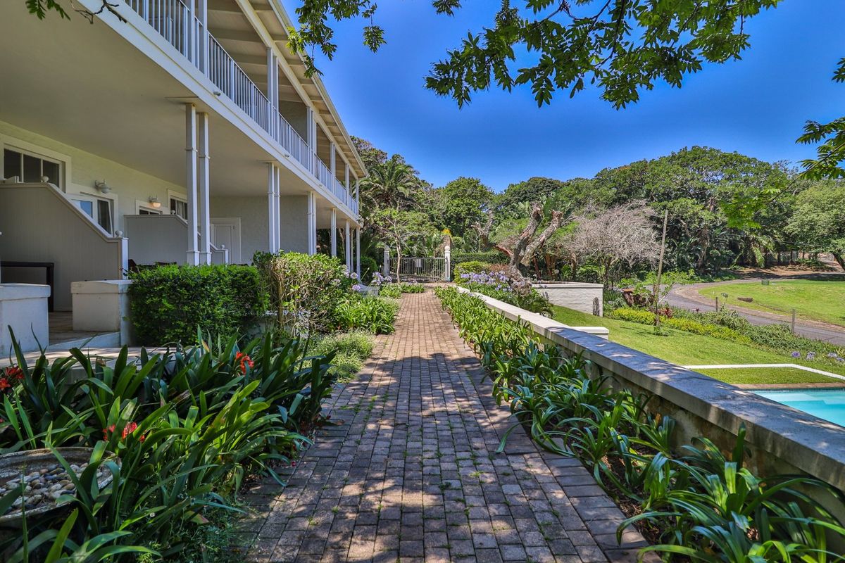A well-maintained exterior walkway featuring brick paving, landscaped garden beds, and a covered veranda along the side of the property.