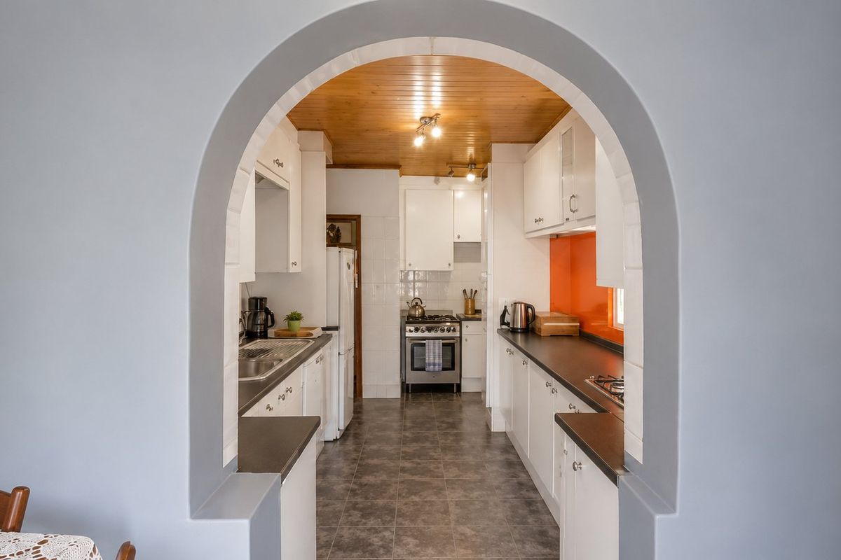 A galley-style kitchen featuring white cabinetry, dark countertops, an orange glass splashback, and a timber-paneled ceiling, viewed through a classic arched doorway.