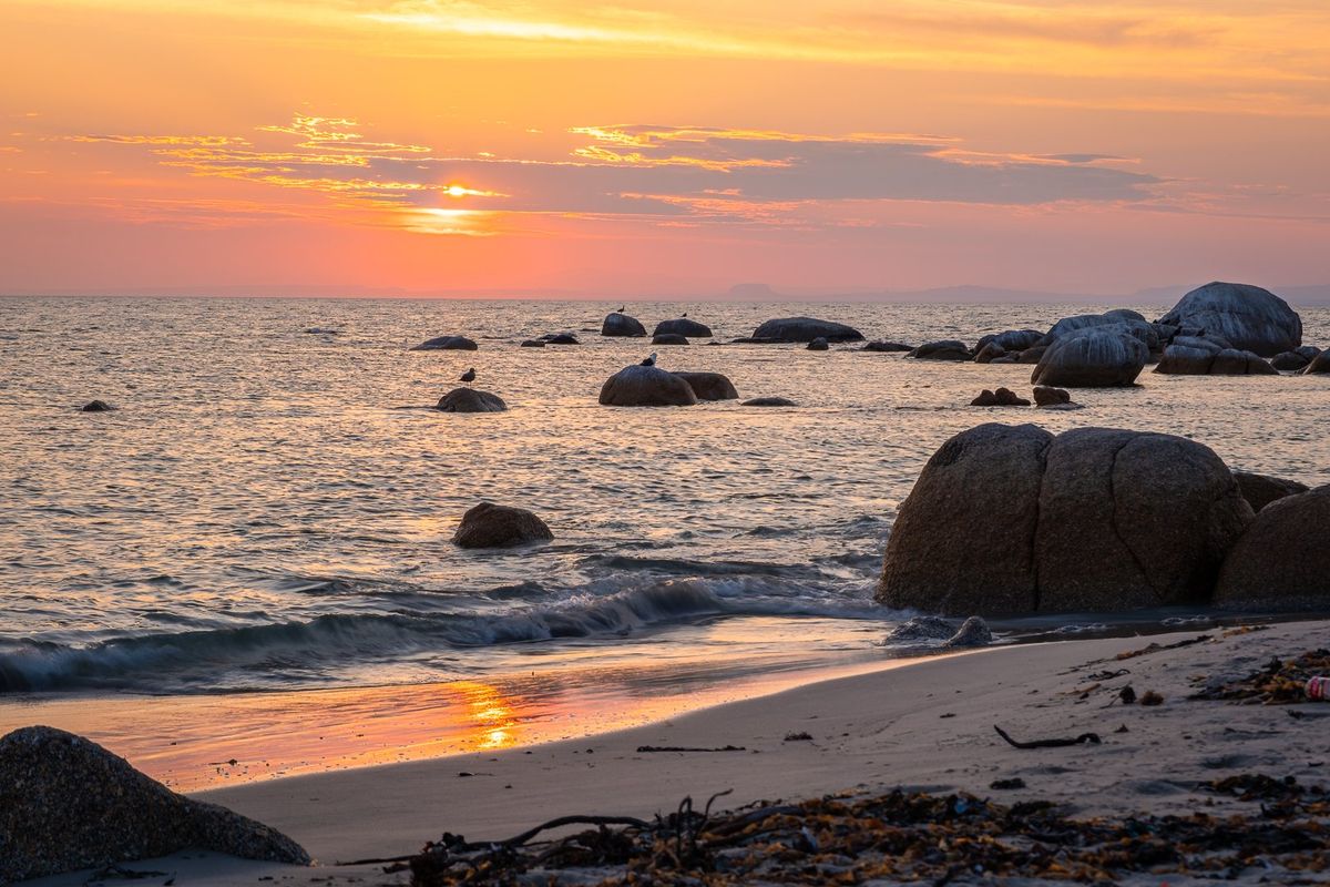 A scenic view of the coastline at sunset, featuring sandy shores, large rock formations, and calm ocean waters.