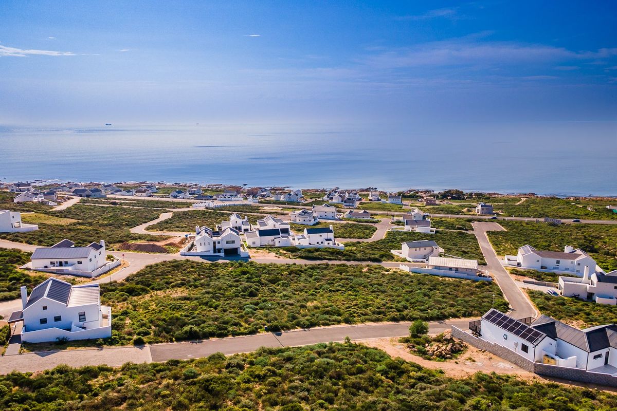 Aerial view of a coastal residential estate featuring modern white homes with dark roofs, paved roads, and proximity to the ocean.