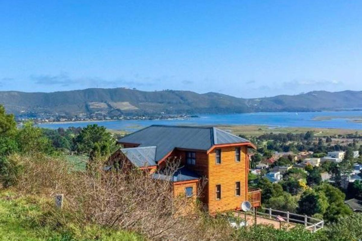 An elevated view of a timber-clad house featuring a metal roof and balcony, set against a scenic landscape with water and mountain views.