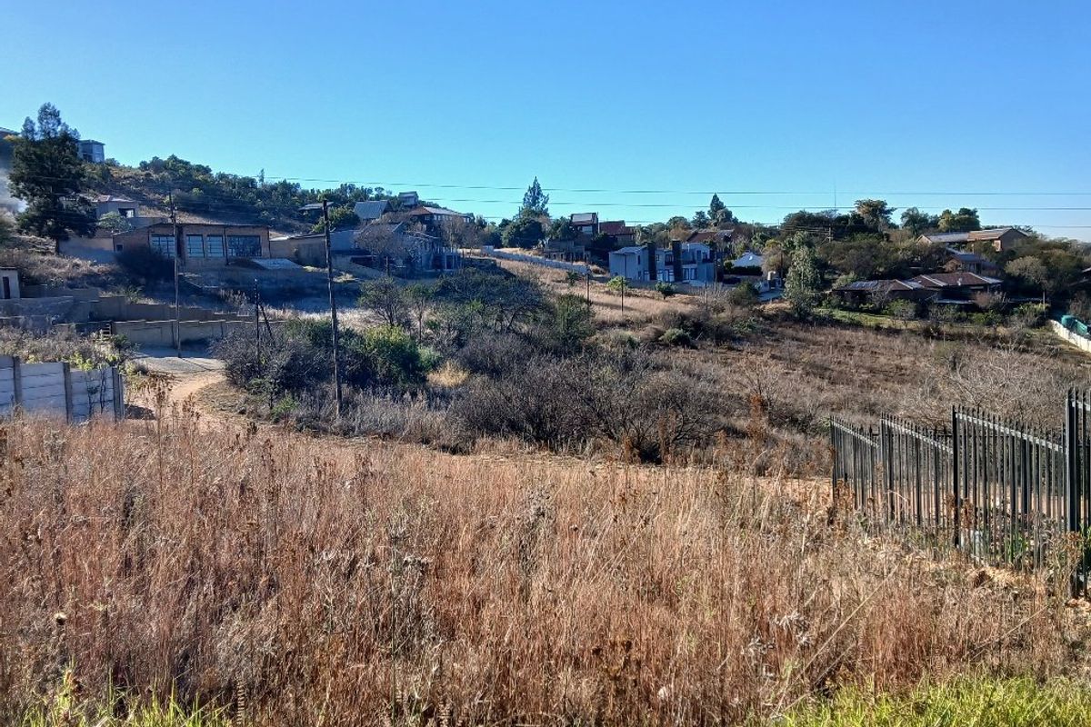 A view of a vacant residential plot featuring natural vegetation, sloping terrain, and surrounding neighborhood housing in the distance.