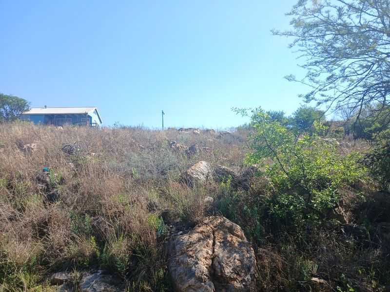 A view of a sloped, undeveloped residential plot featuring natural rocky terrain and native vegetation with a neighboring structure visible in the distance.