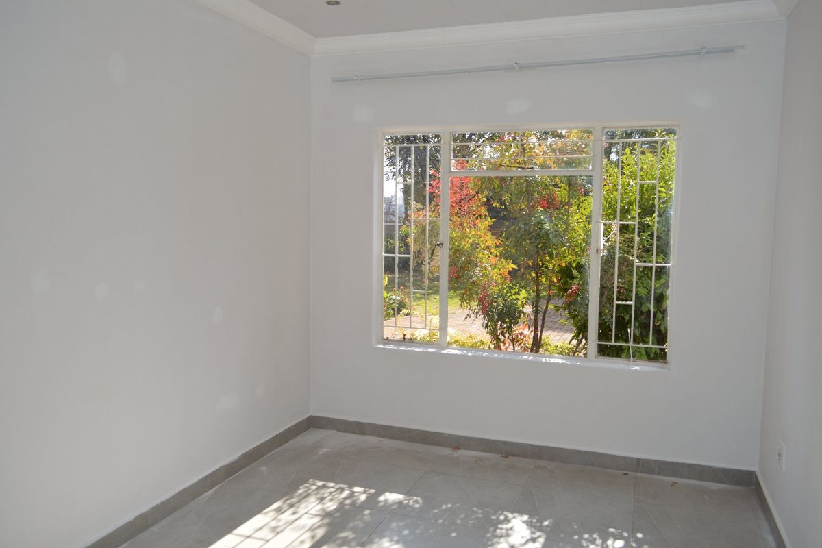 A bright, empty room featuring tiled flooring, white walls with visible patch work, and a large window with security bars overlooking the garden.