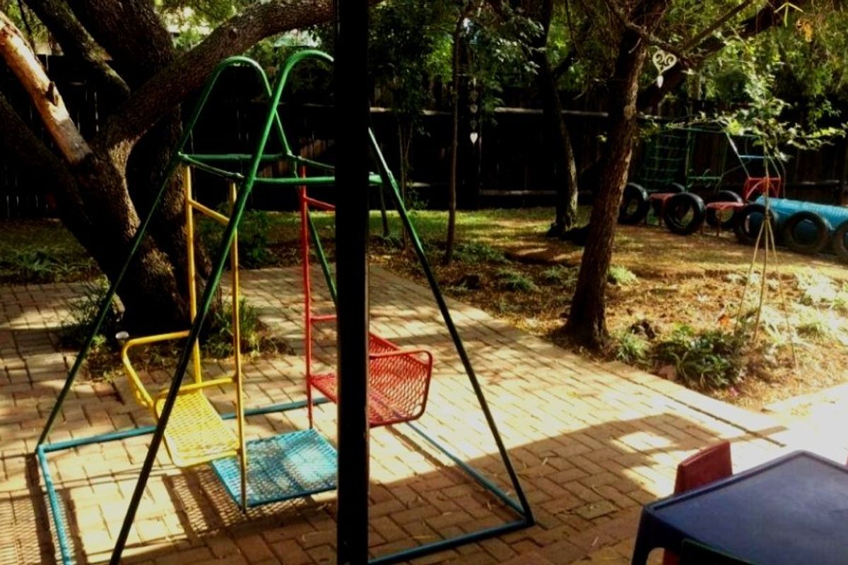 A shaded backyard garden area featuring paved flooring, mature trees, and a dedicated play space with fixed metal playground equipment.