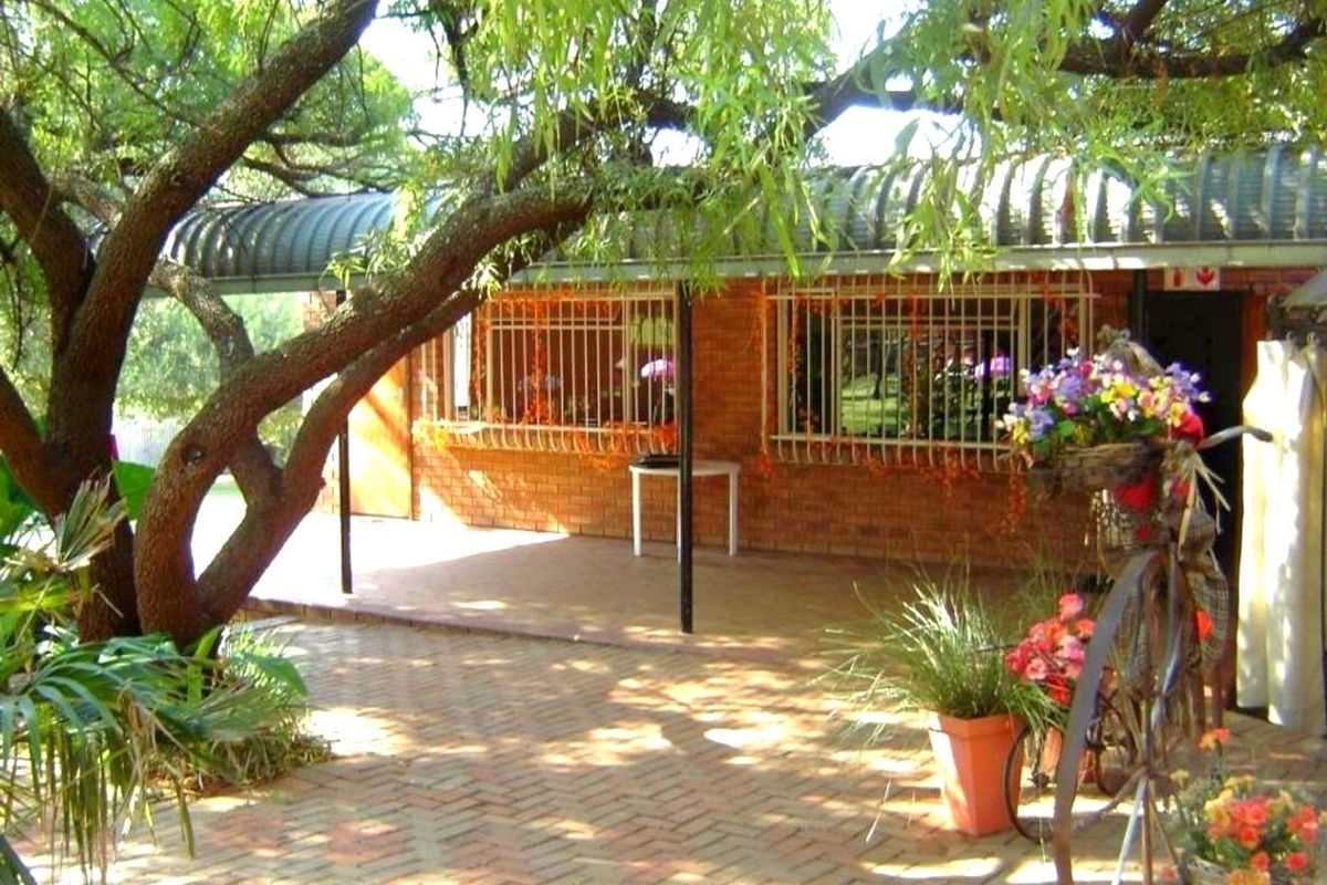 A covered patio area featuring brick paving, face brick exterior walls, and security bars on the windows, shaded by mature trees.