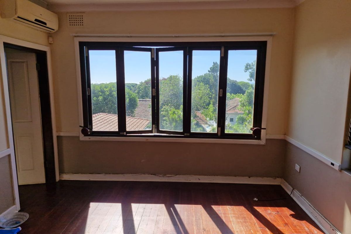 A bright, empty bedroom featuring polished timber flooring, a large multi-pane window with garden views, and a wall-mounted air conditioning unit.