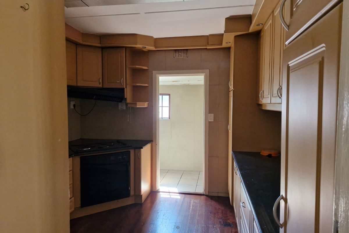 A dated kitchen featuring built-in wooden cabinetry, dark countertops, and an integrated oven and hob, leading into an adjacent utility space.