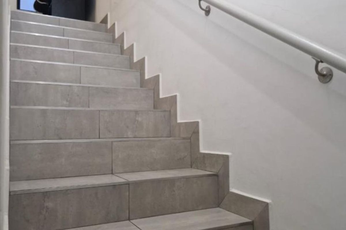 A staircase featuring modern grey tiled steps and a wall-mounted handrail, leading to an upper landing area.